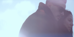 Cinematic music video production shot of a romantic couple embracing on a Parisian bridge with a bright blue sky background.
