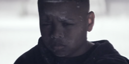 Cinematic music video production shot of a focused boy in a hoodie sitting in a gritty, atmospheric stairwell.