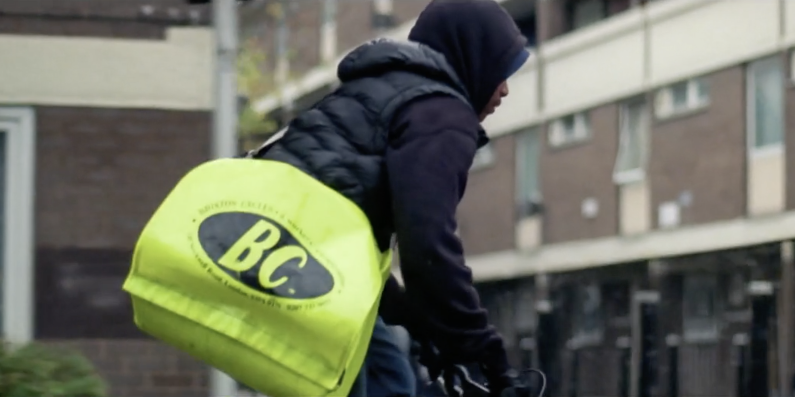 Teenage Paper Boy In Hoodie And Body Warmer Riding BMX Through South London With Council Estate Background