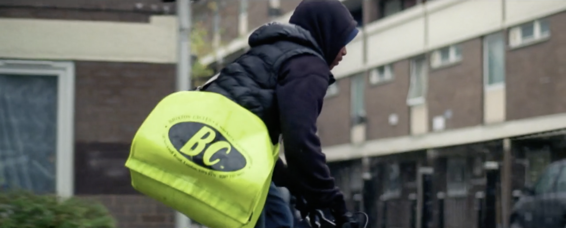 Teenage Paper Boy In Hoodie And Body Warmer Riding BMX Through South London With Council Estate Background