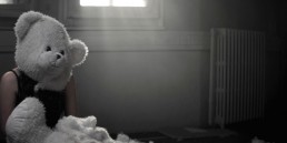 Cinematic music video production shot featuring a surreal, dark narrative scene with a woman in a teddy bear mask in a derelict room.