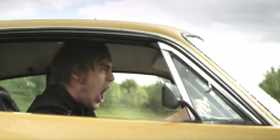 Cinematic music video production still featuring an intense narrative driving scene with a man in a yellow American muscle car.