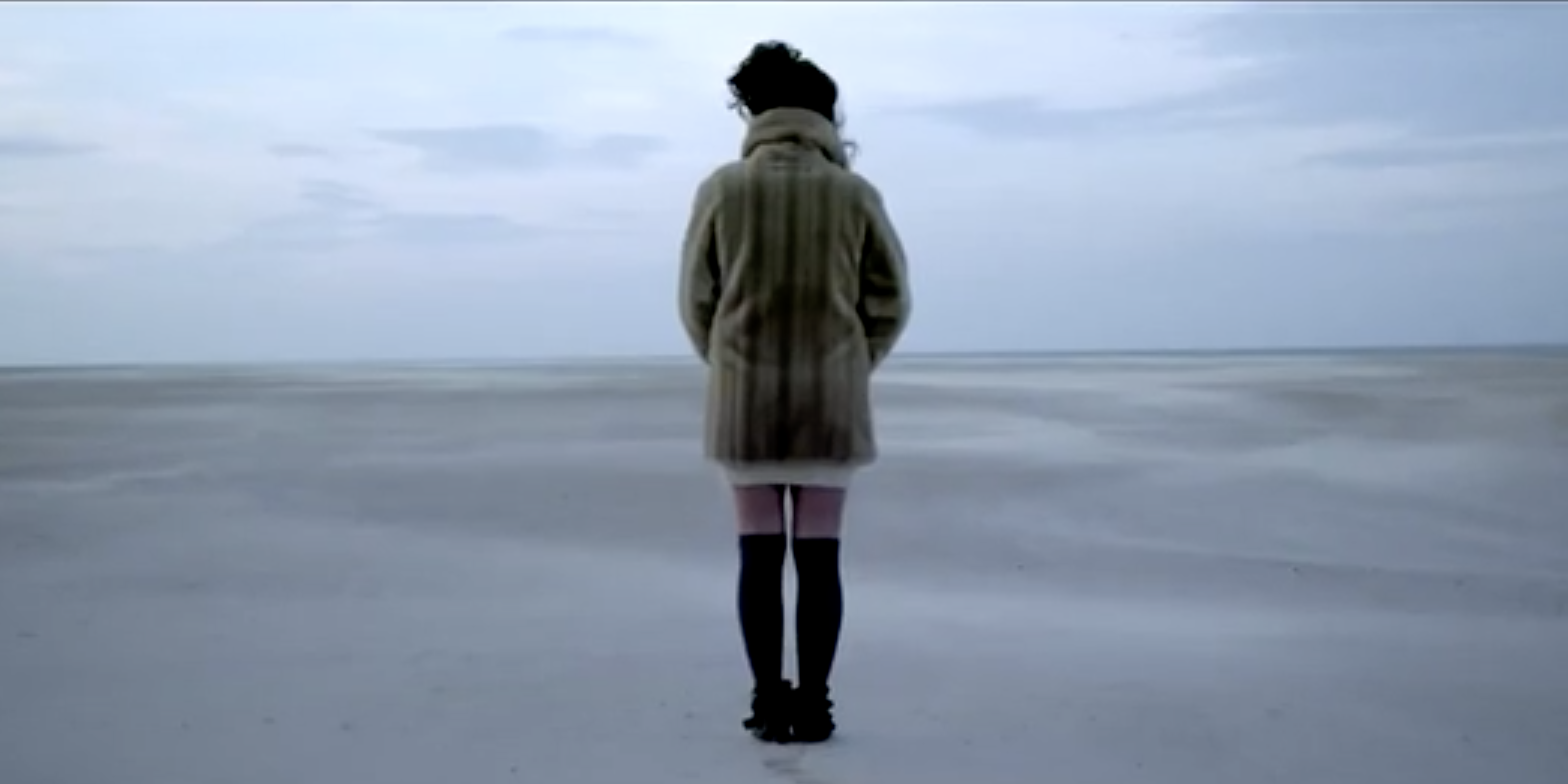 Female Singer In Fur Coat Standing On Sandy Beach With Ocean Background