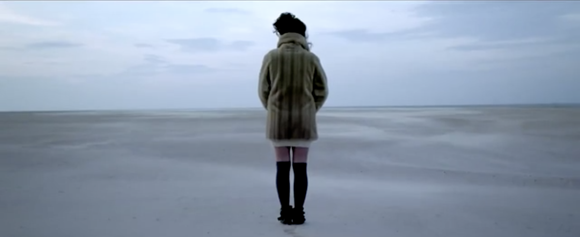 Female Singer In Fur Coat Standing On Sandy Beach With Ocean Background