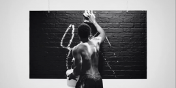 Cinematic music video production shot featuring a stylized performance scene of a man covered in white paint against a black brick wall.