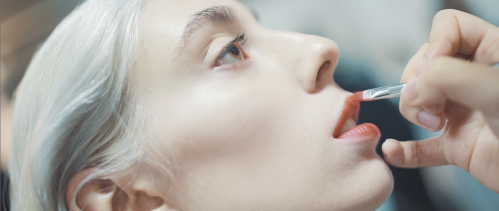 Beautiful Female Model With Brown Eyes And Grey Hair Having Lipstick Applied By Makeup Artist At Fashion Runway Show