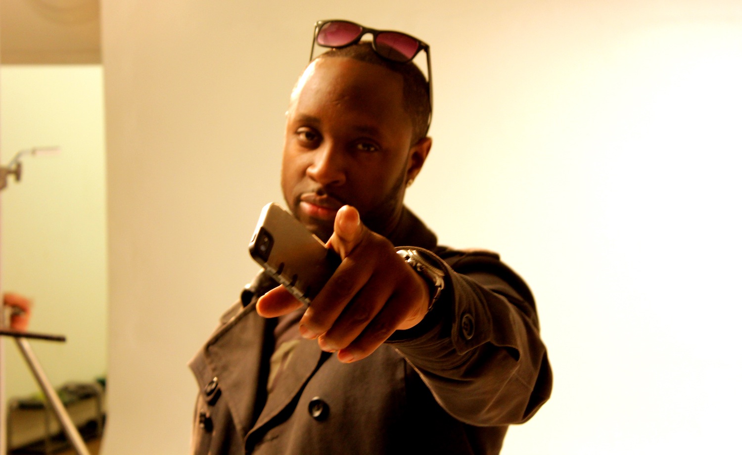 Professional Photography Closeup Of Black Man In Khaki Jacket Wearing Sunglasses Holding Phone Pointing To Camera On White Studio Backdrop