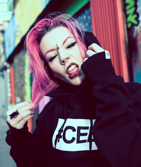 Professional Photography White Woman With Pink Hair Wearing Black Hashtagcee Hoody With White Logo In Front Of Shop Shutters