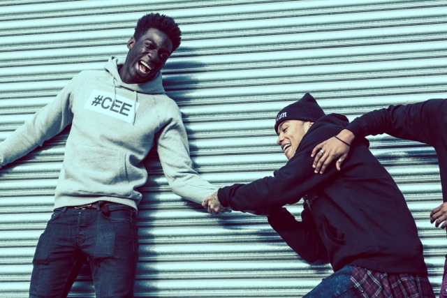 Professional Photography Black Man Wearing Grey Hashtagcee Hoodie Being Pulled By Man Wearing Black Hashtagcee Generals Hoody In Front Of Grey Shutters