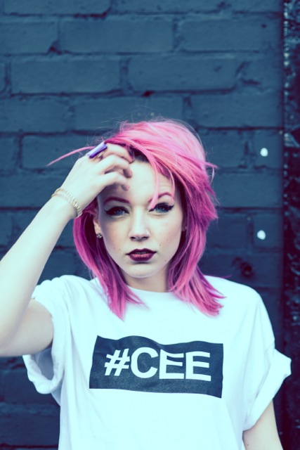 Professional Photography White Woman With Pink Hair Standing In Front Of Black Brick Wall Wearing White Hashtagcee T-Shirt