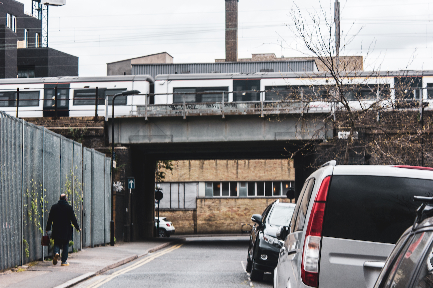 Professional Photography Man With Bag And Black Coat About To Walk Under Bridge With Train Passing Above In London Fields Hackney East London