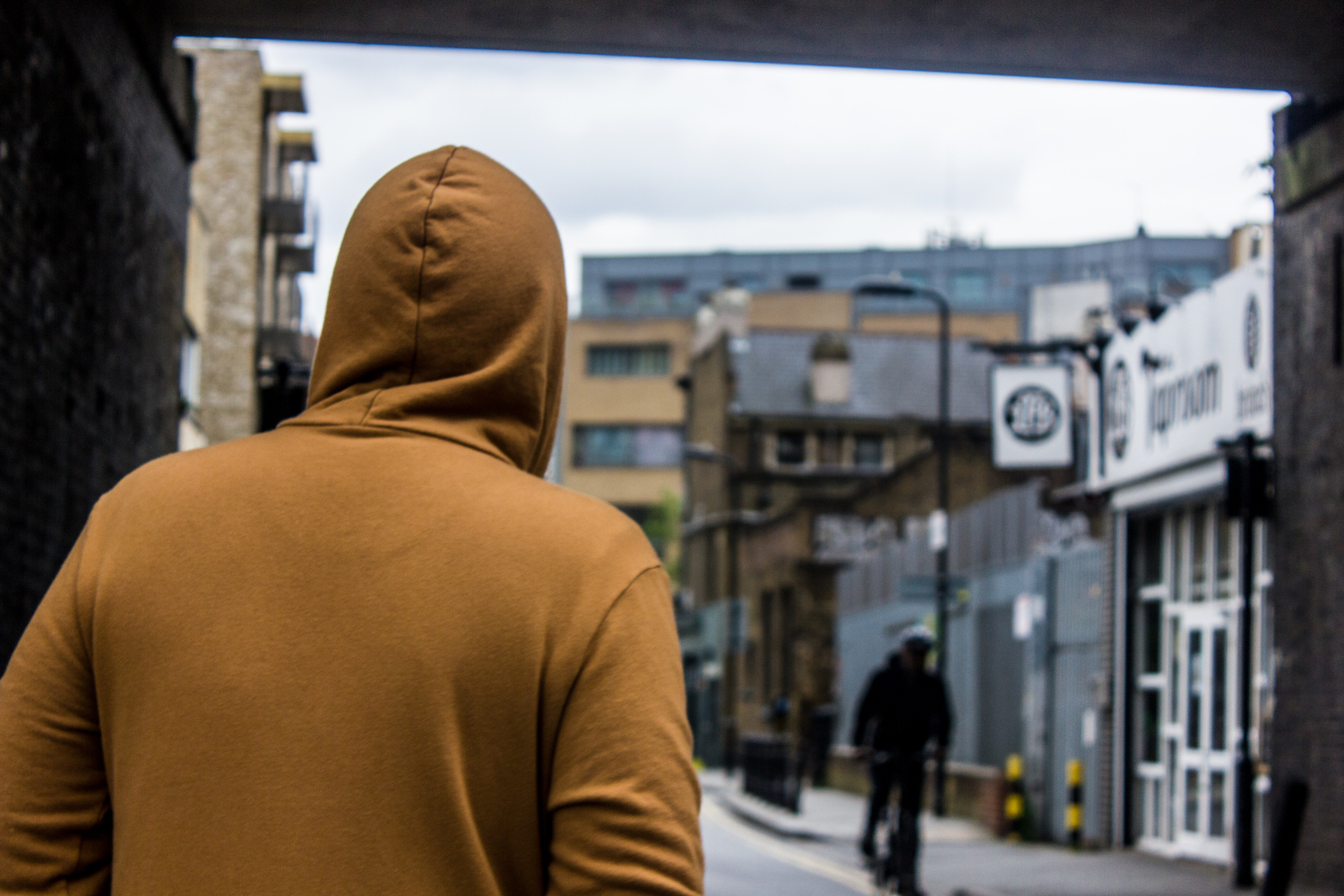 Professional Photography Man In Orange Hoodie Walking Under Dark Bridge As Cyclist Approaches In London Fields Hackney