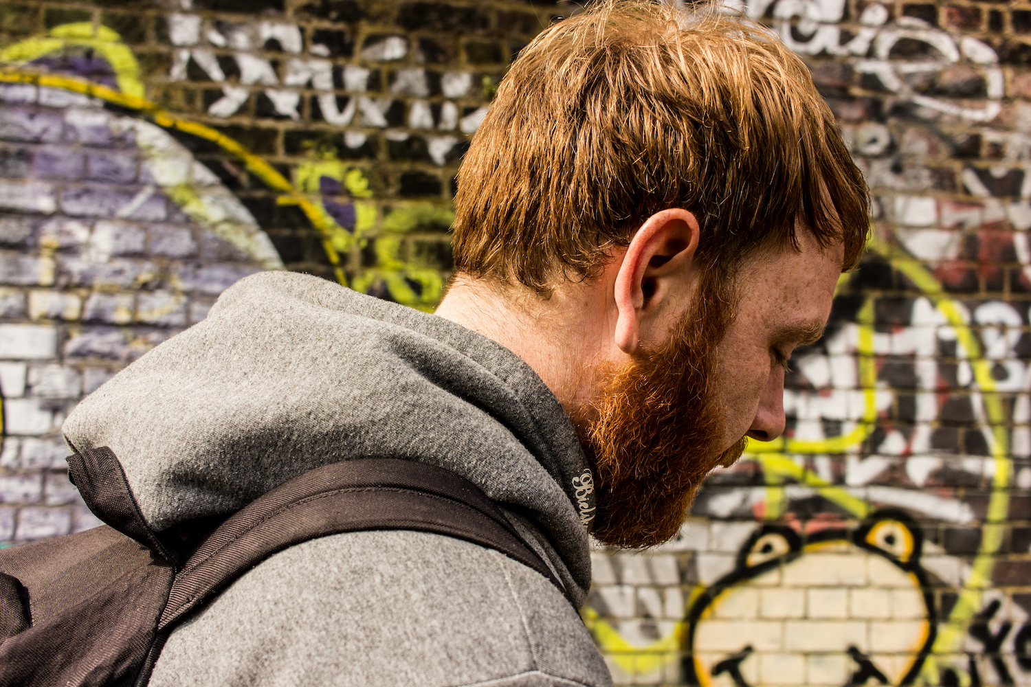 Professional Photography White Man With Beard Grey Hoodie And Black Backpack Walking In Front Of Graffiti Wall