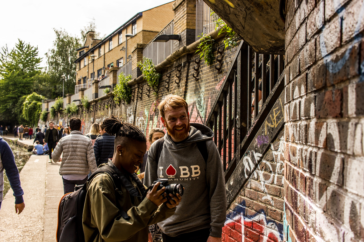 Professional Photography White Man With Beard In Grey Hoody Black Jeans And Backpack With Black Woman Wearing Khaki Jacket Dreadlocks And Black Backpack Filming Sign With Camera Next To Canal River