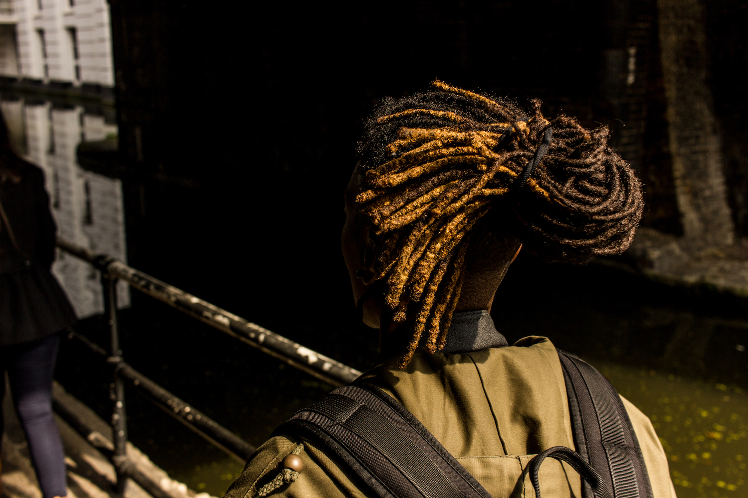 Professional Photography Black Woman With Khaki Jacket Black Backpack And Dreadlocks Walking Under Bridge In Little Venice Paddington London