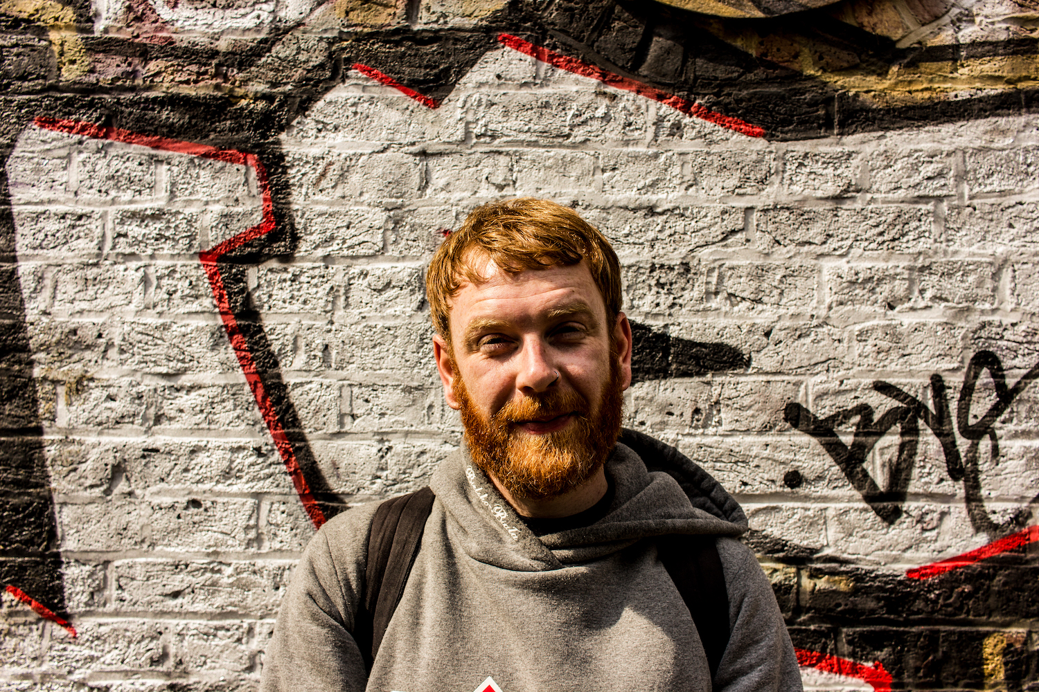 Professional Photography White Man With Beard Grey Hoody And Black Backpack Standing In front of Graffiti Wall