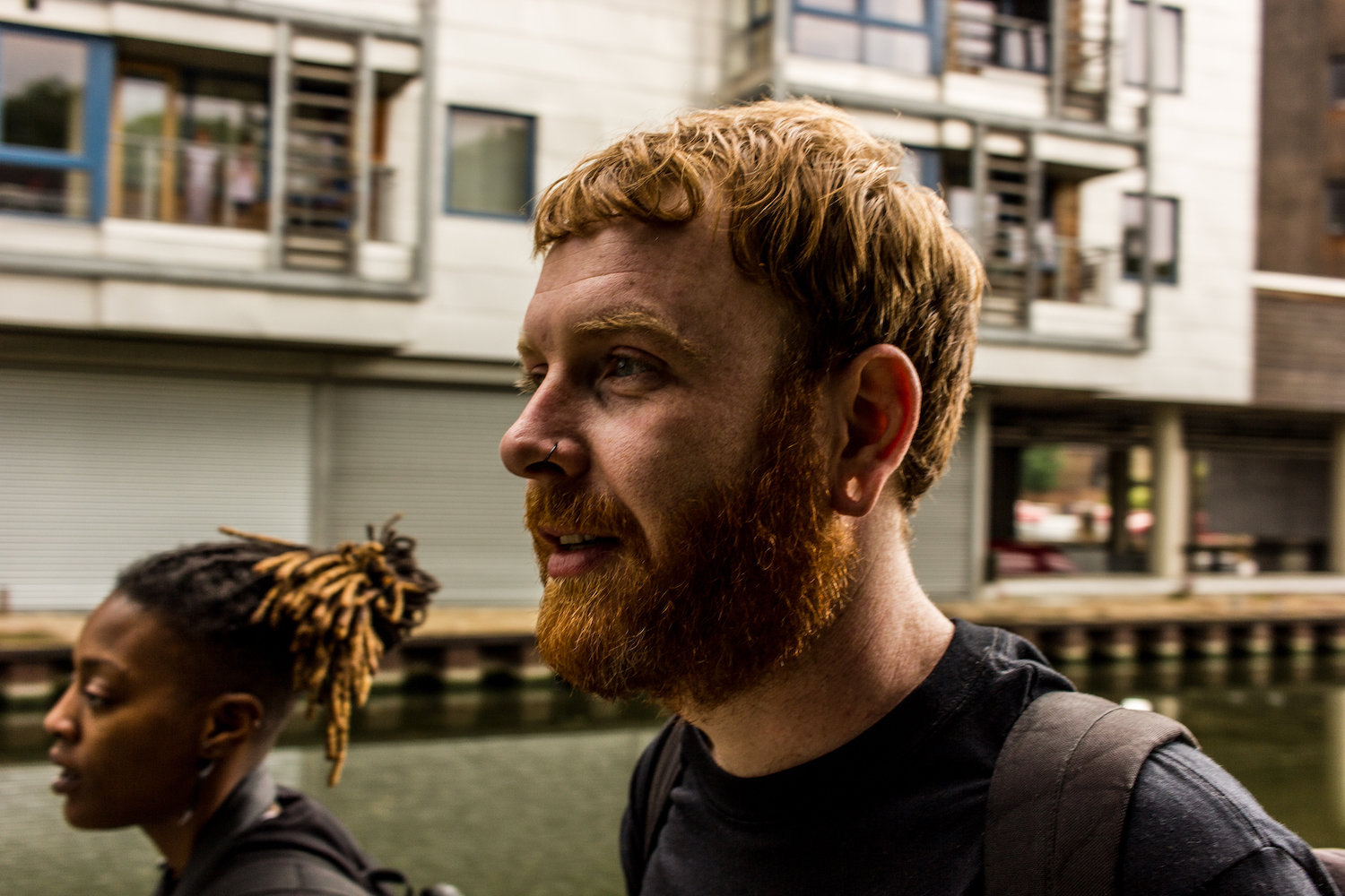 Professional Photography White Man With Beard Black T-Shirt And Black Backpack Walking With Dreadlocks Black Woman Carrying Black Backpack Buildings And River Canal In Background Camden London