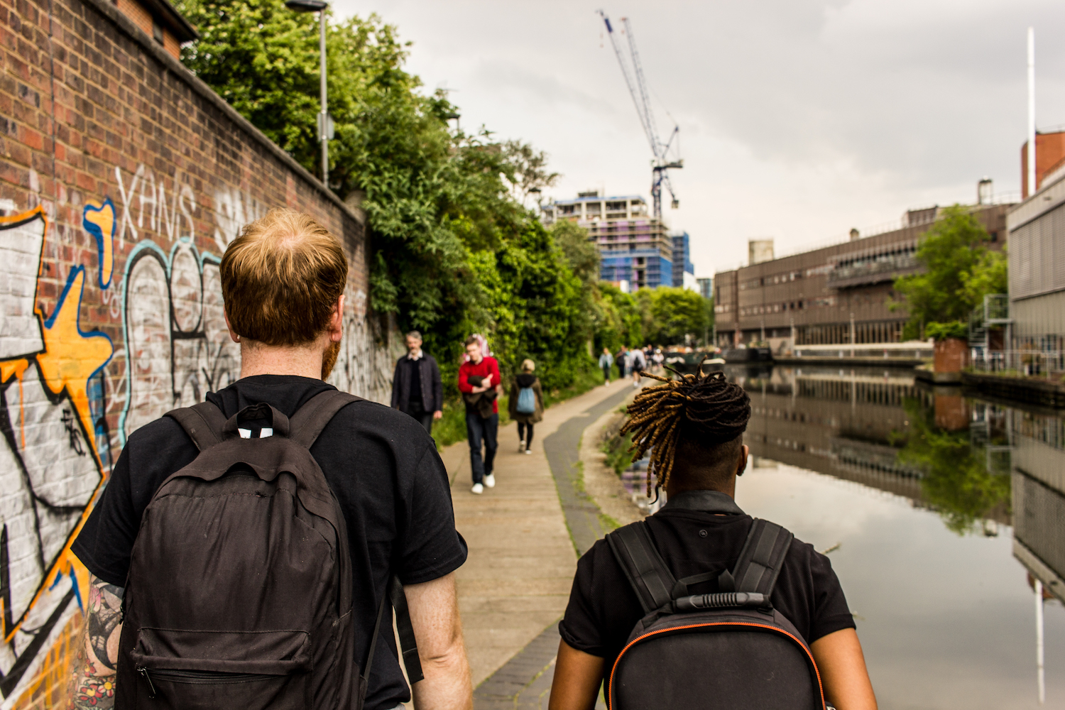 Professional Photography White Man With Beard Black T-Shirt And Black Backpack Walking With Dreadlocks Black Woman Carrying Black Backpack Walking Alongside River Canal In Camden With Graffiti Crane And Buildings In Background