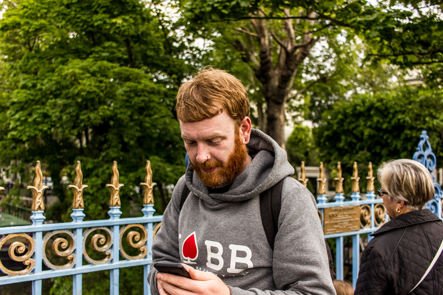 Professional Photography White Man Beginning Ten Mile Charity Walk Wearing Grey Hoody Standing Looking At Phone With Green Trees In Little Venice Paddington London