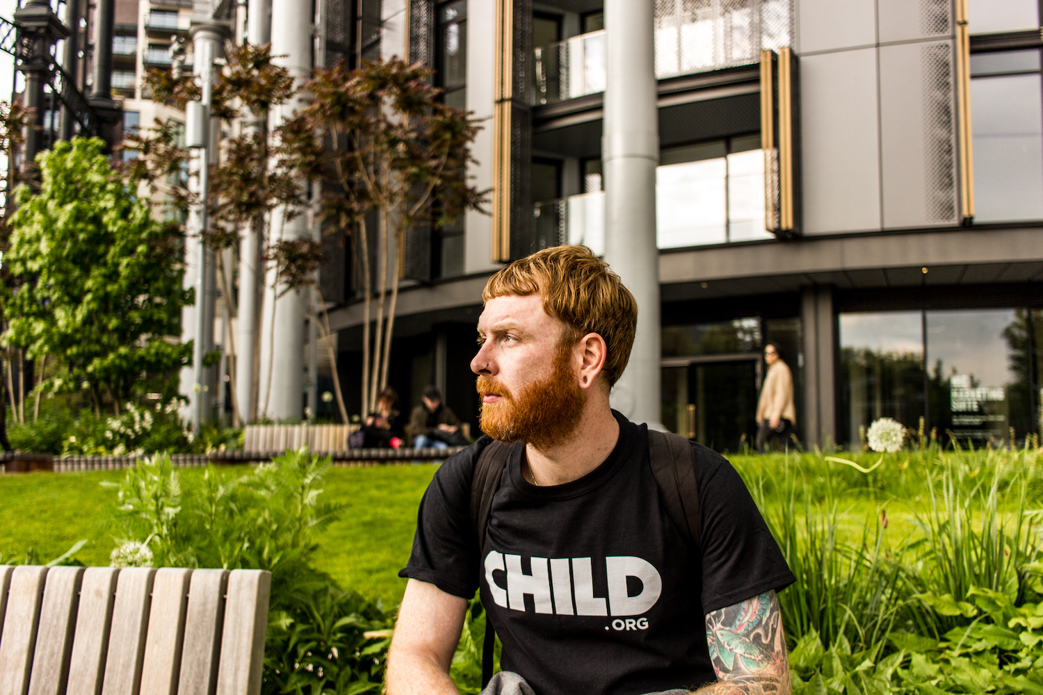 Professional Photography White Man With Beard Wearing Black Child.org T-Shirt Sitting On Bench In Front Of Bushes And Metal Buildings In Camden London