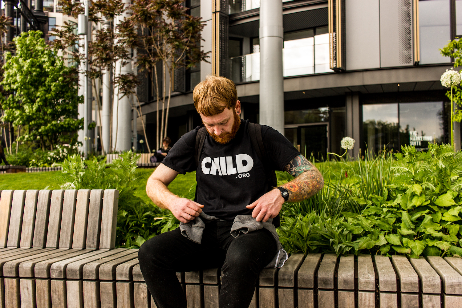 Professional Photography White Man With Beard Showing Black Child.org T-Shirt Sitting On Bench In Front Of Bushes And Metal Buildings In Camden London