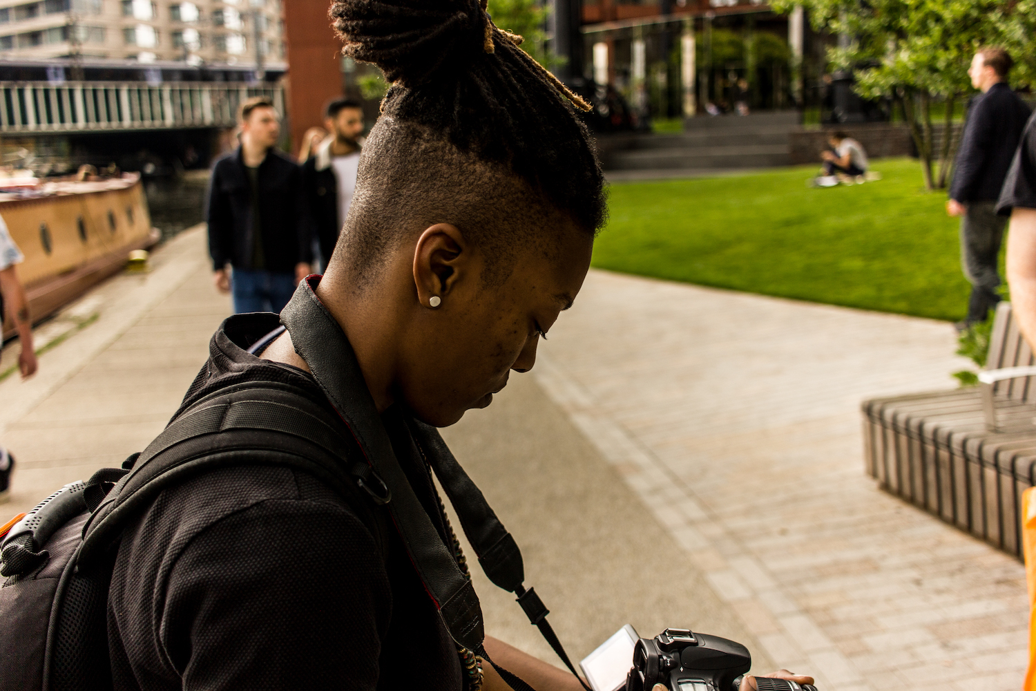 Professional Photography Black Woman Wearing Black T-Shirt With Black Backpack And Dreadlocks Filming With Camera Green Grass And People In Background Camden London