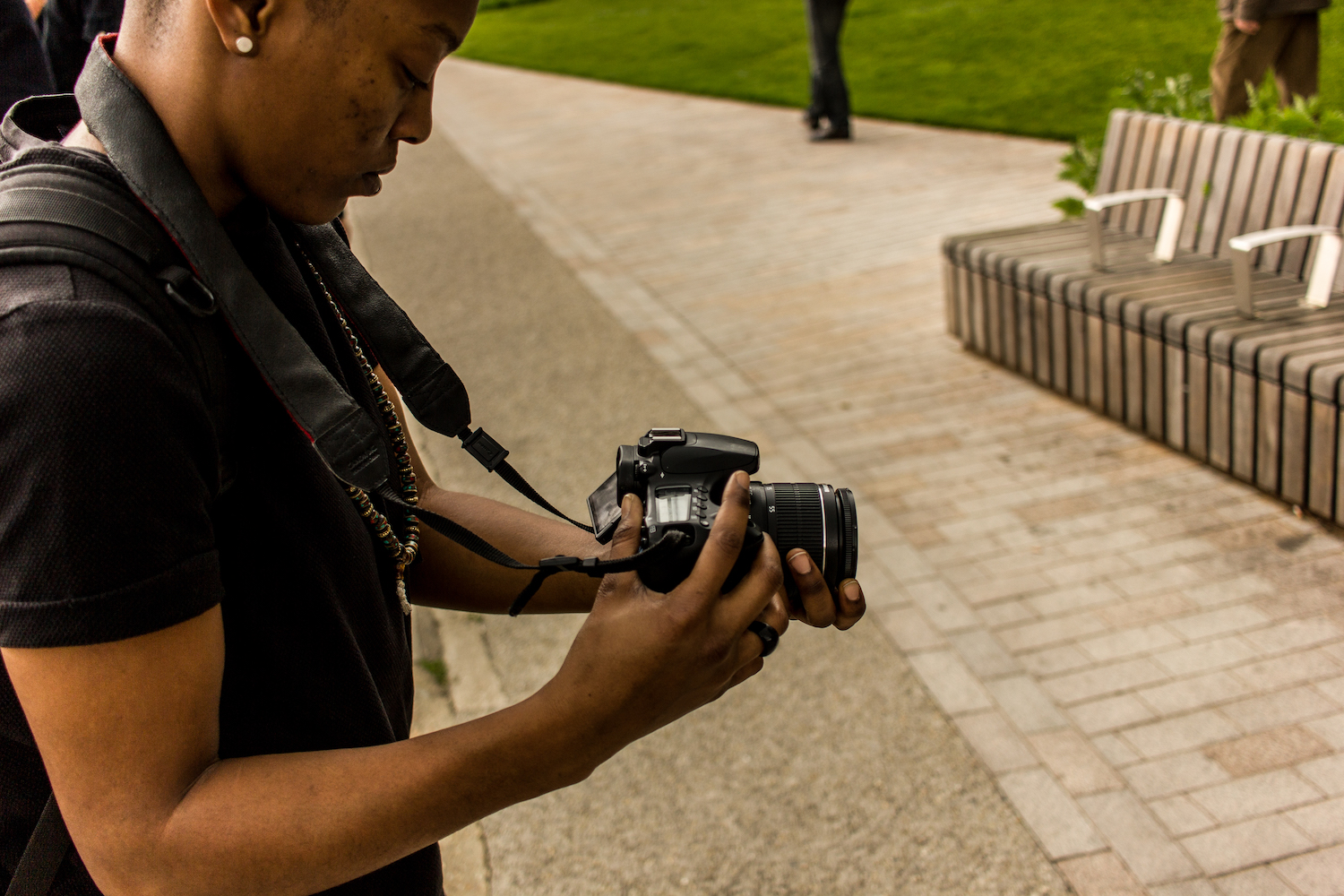 Professional Photography Close-Up Of Black Woman Wearing Black T-Shirt With Black Backpack And Dreadlocks Filming With Camera Green Grass In Background Camden London