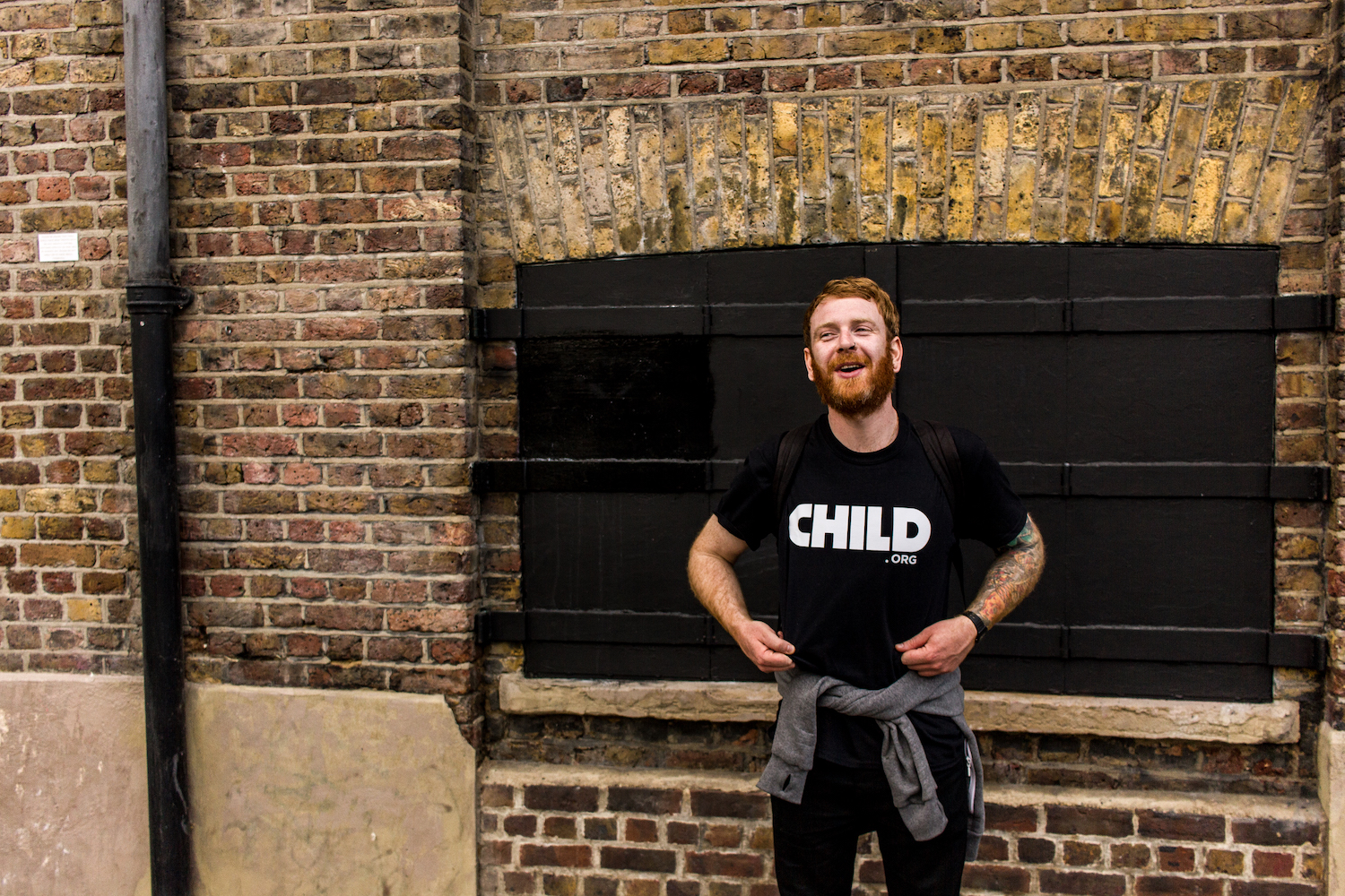 Professional Photography White Man With Beard Showing Black Child.org T-Shirt Standing In Front Of Brick Wall In Camden