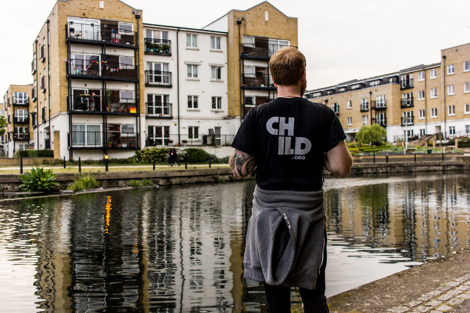 Professional Photography White Man Wearing Black Child.org T-Shirt And Grey Hoody Wrapped Around Waist Standing In Front Of River Canal With Buildings In Background