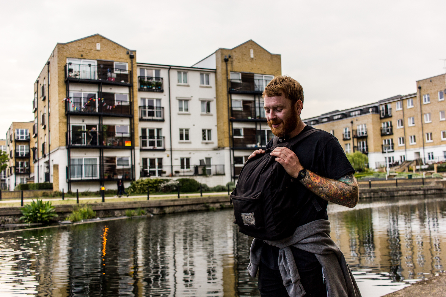 Professional Photography White Man With Beard Wearing Black T-Shirt Grey Hoody Around Waist Holding Black Backpack Over Stomach In Front Of River Canal With Buildings In Background