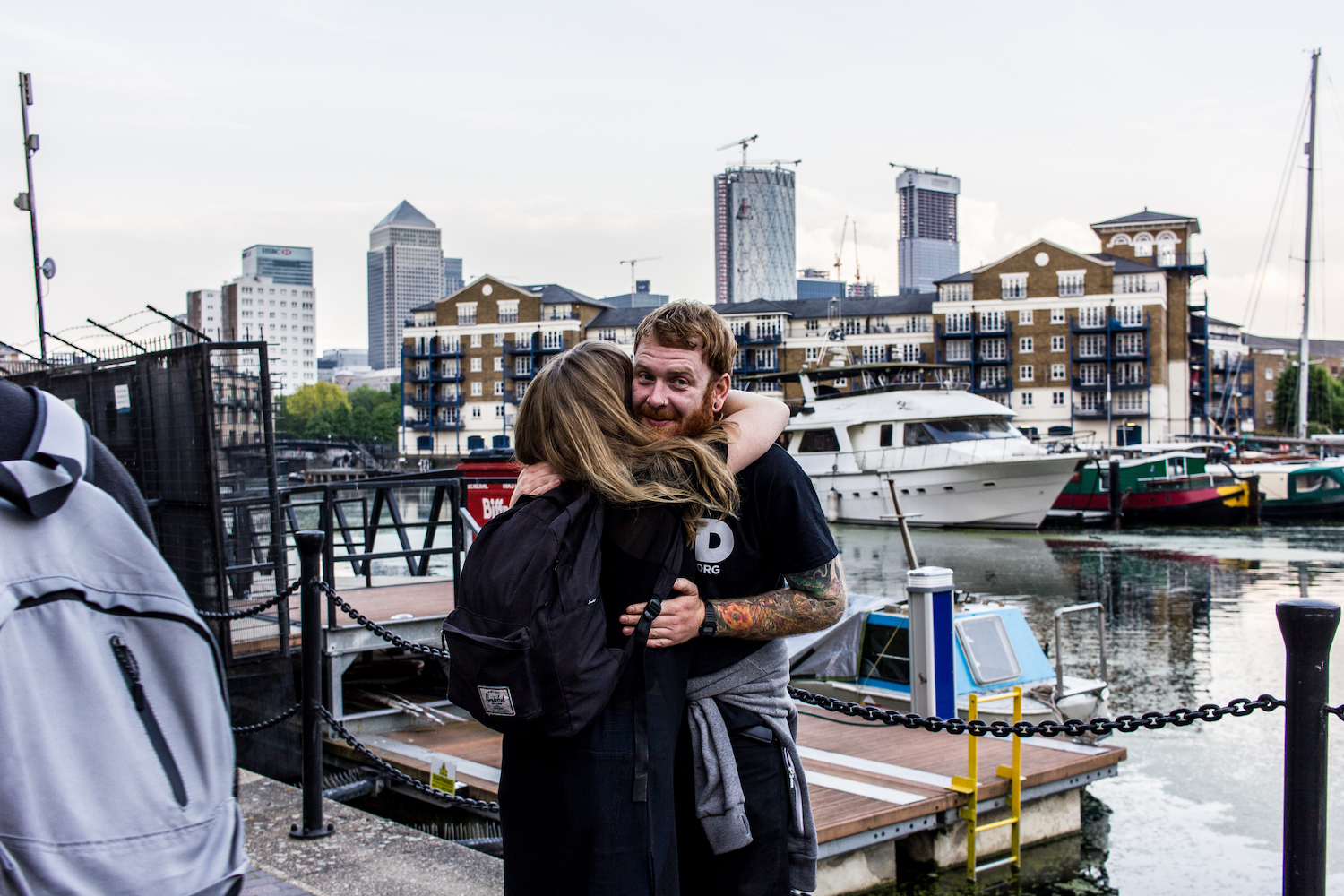 Professional Photography White Man With Beard Wearing Black T-Shirt And Grey Hoody Around Waist Hugging Woman In Limehouse Basin With Boats River Canal And Canary Wharf Buildings In Background