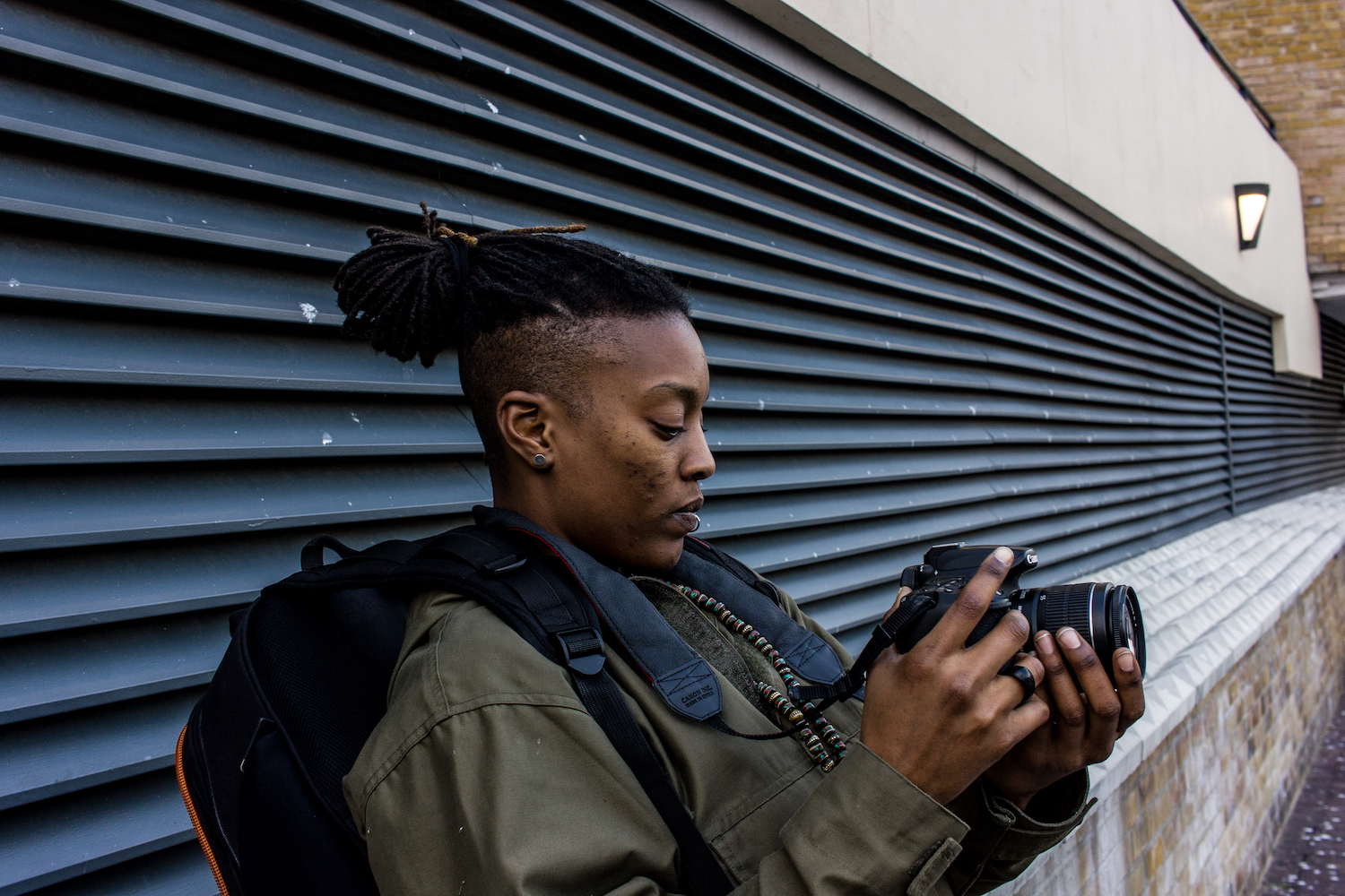 Professional Photography Black Woman With Lip Ring Khaki Jacket Black Backpack And Dreadlocks Leaning Against Wall With Vents Filming With Camera In Limehouse Basin