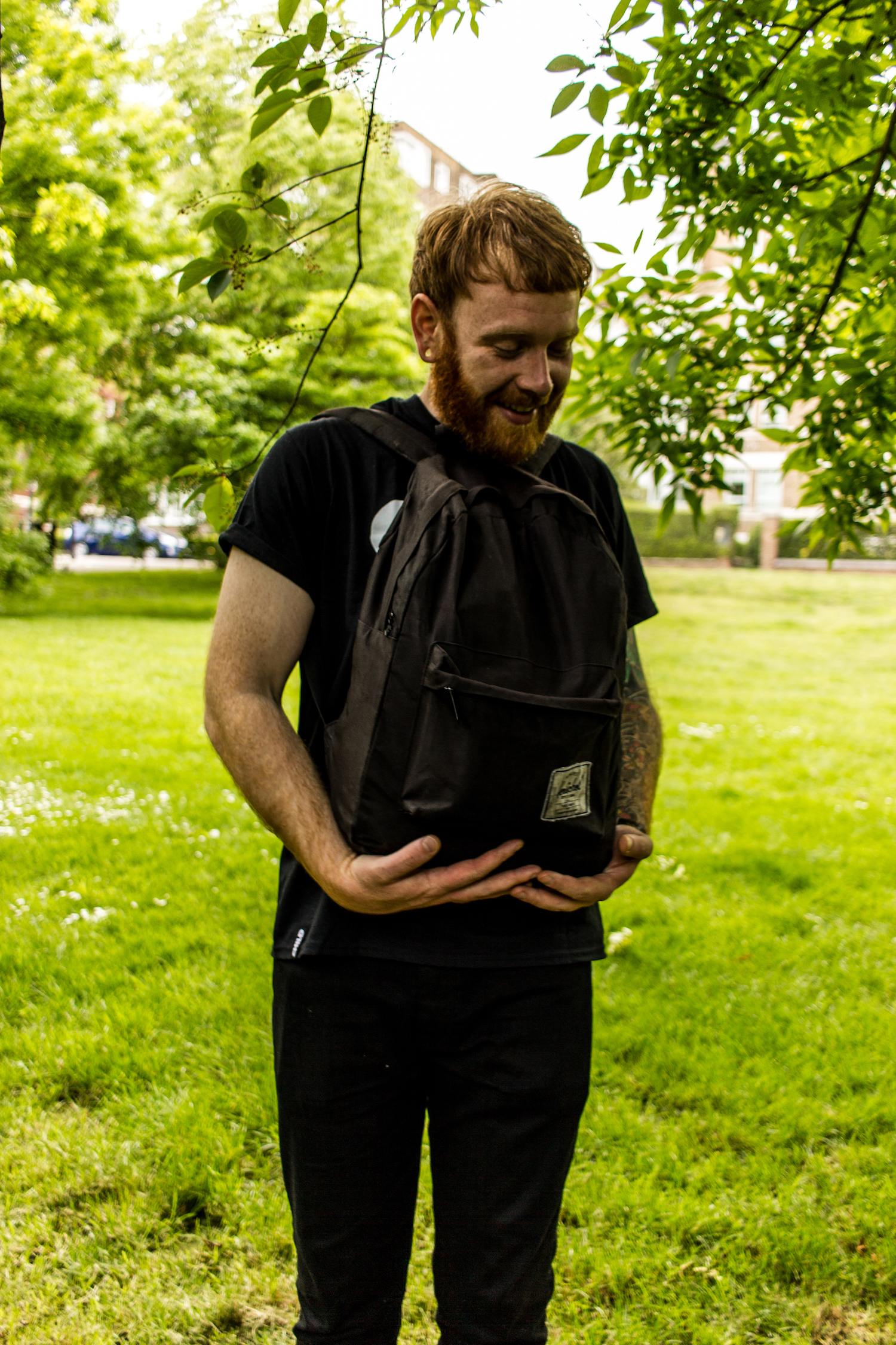 Professional Photography White Man With Beard Black T-Shirt And Black Jeans Standing Holding Black Backpack With Green Grass And Trees In Background In Little Venice Paddington London