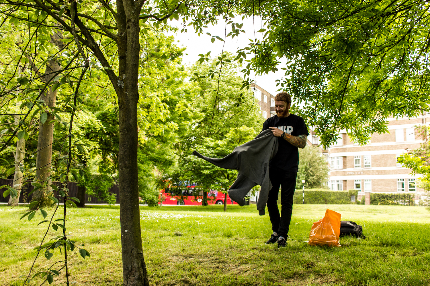 Professional Photography White Man With Beard Black T-Shirt And Black Jeans Holding Grey Hoody With Green Grass Trees And Buildings In Background In Little Venice Paddington London