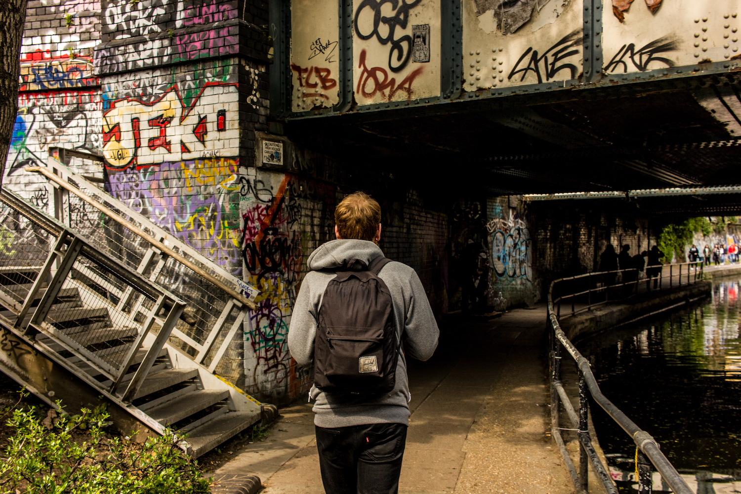 Professional Photography White Man In Grey Hoody Black Jeans And Backpack Walking Under Bridge With Graffiti Next To Canal River In Little Venice Paddington London