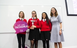 Professional Photography Three Award Winning Female Students Wearing Red Sweatshirts Holding Silver And Pink Trophy At Event With Woman In Grey Dress
