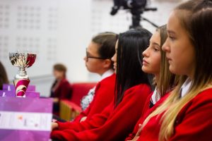 Professional Photography Four Female Students Wearing Red Sweatshirts Sitting Watching Event With Silver And Pink Trophy