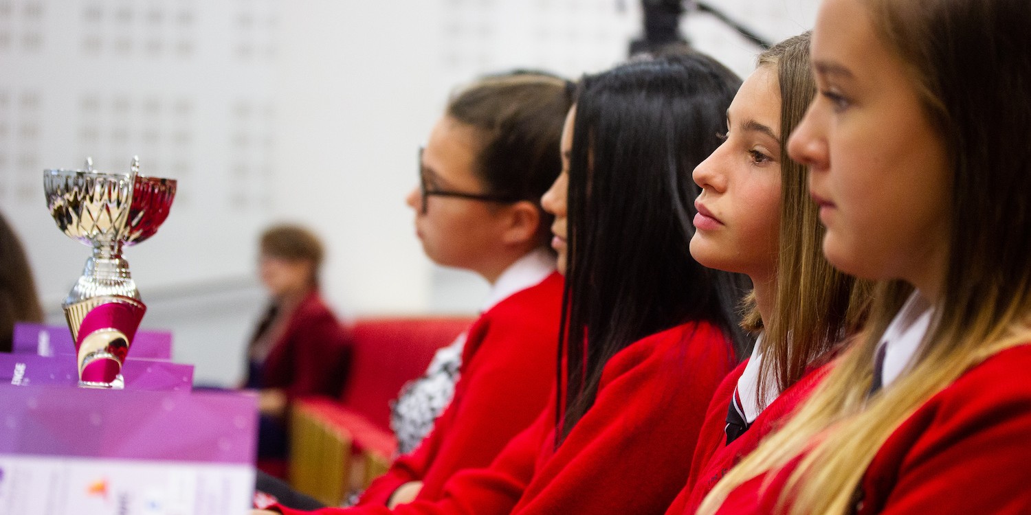 Professional Photography Four Female Students Wearing Red Sweatshirts Sitting Watching Event With Silver And Pink Trophy