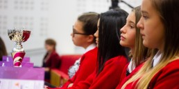 Professional Photography Four Female Students Wearing Red Sweatshirts Sitting Watching Event With Silver And Pink Trophy