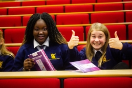 Professional Photography Two Female Students Sitting On Red Seats Wearing Blue Sweatshirts Smiling With Thumbs Up.
