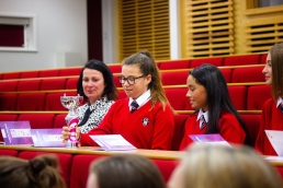 Professional Photography Three Female Students Wearing Red Sweatshirts Sitting In Row At Event Admiring Trophy With Woman In Multicolour Shirt