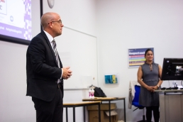 Professional Photography Man Wearing Navy Blue Suite White Shirt And Striped Tie Stands Hands Clasped Addressing Students At Event With Woman In Background